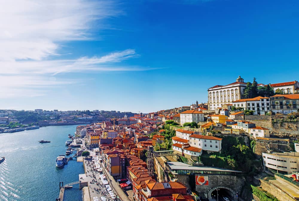 Casas em um morro com um rio à esquerda, com pessoas em uma calçada e barcos atracados